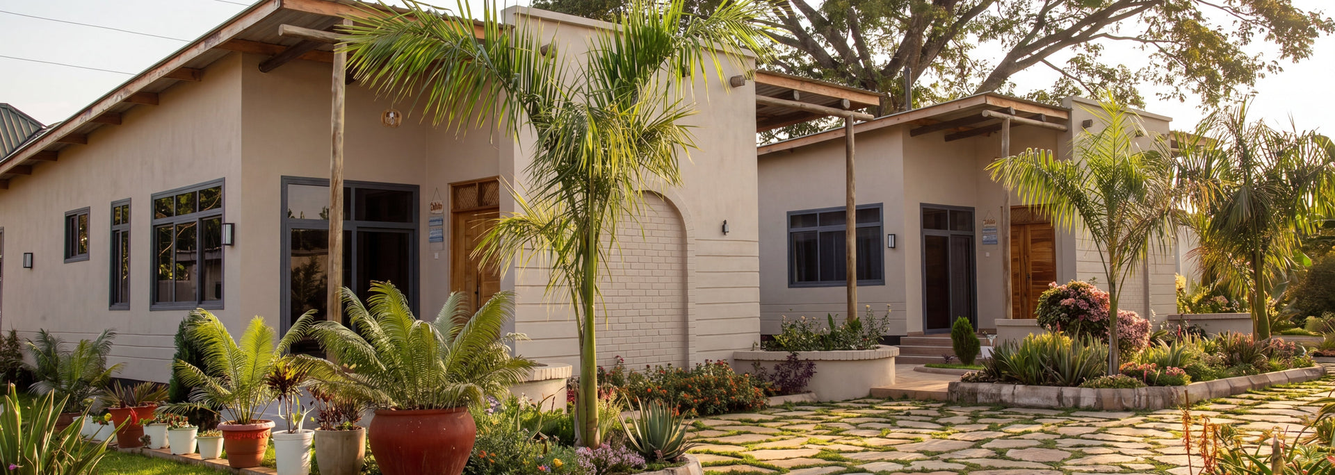 A modern single-story villa with cream-colored stucco walls, dark-framed windows, and a stone pathway leading through a landscaped garden with palm trees and potted plants under a large tree.