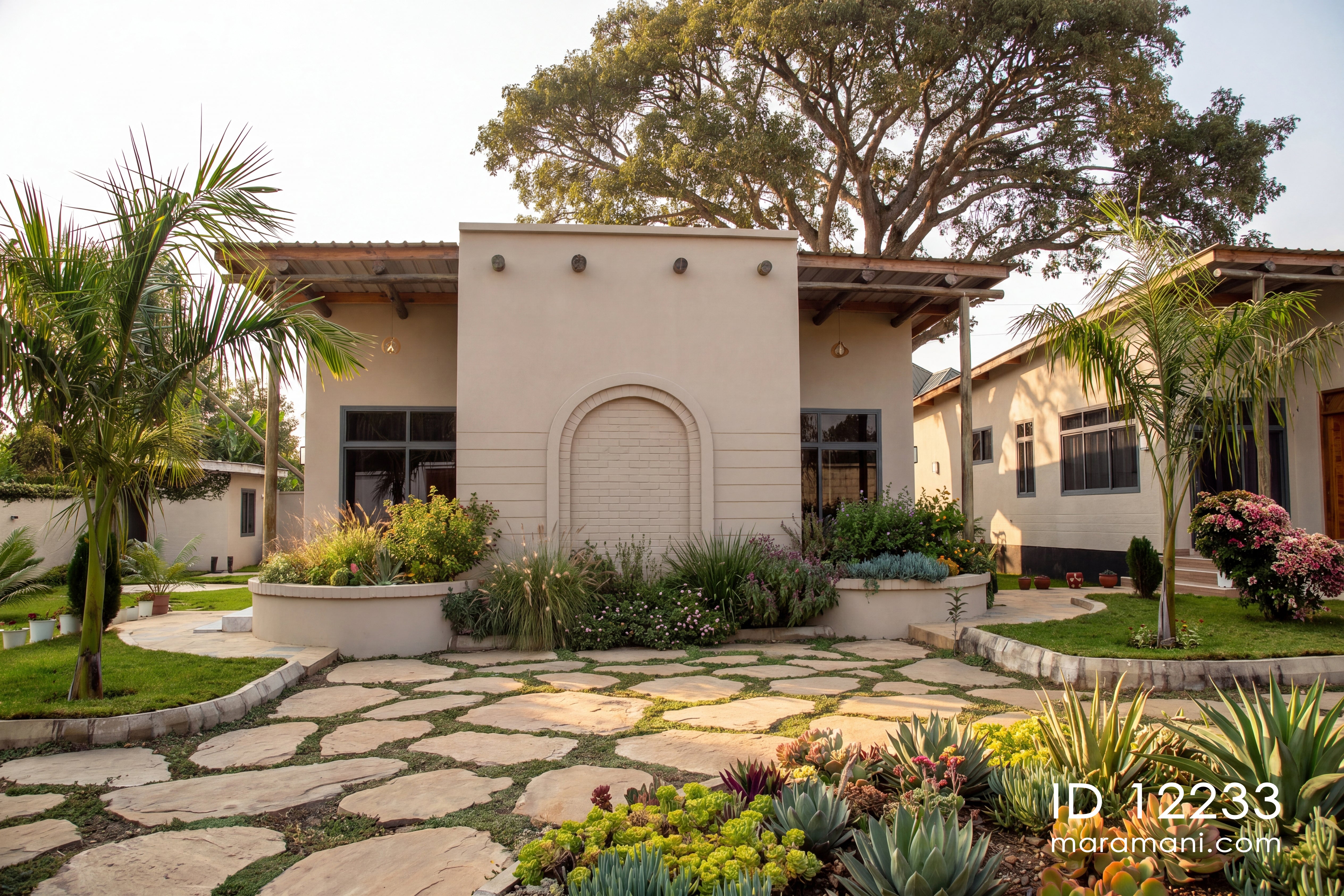 A wide-angle view of a modern single-story stucco house with a large stone patio in the foreground, surrounded by lush landscaping, palm trees, and a tall tree overhead.